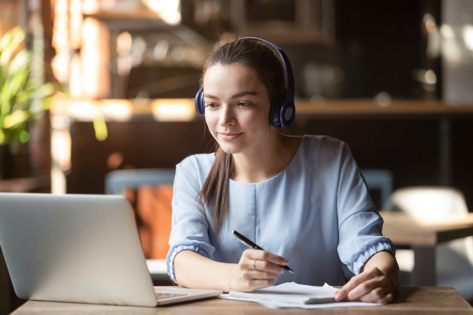Focused woman wearing headphones using laptop, writing notes | Schools Week