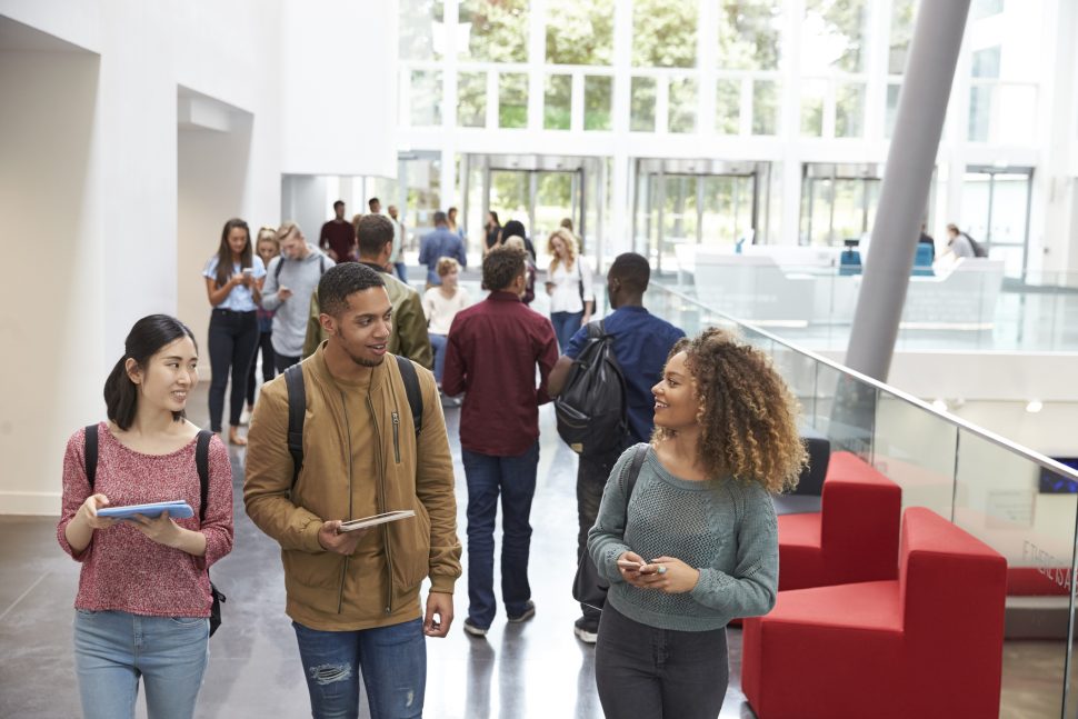 Students holding tablets and phone talk in university lobby | Schools Week Ofqual responded to the DfE's post-qualification admissions (PQA) reforms to university applications.