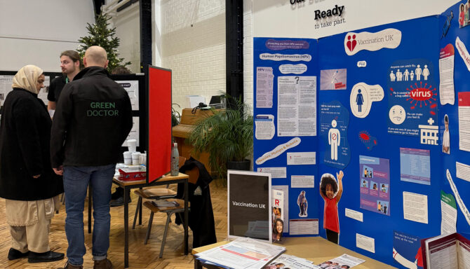 Group of people talking near a vaccine information booth with a large blue wall display labeled Vaccination UK; indoor event setting with posters and chairs nearby.