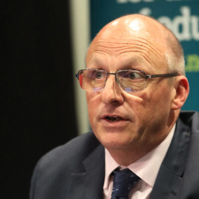 Close-up of a bald man in a dark suit and glasses speaking at a panel, with a blue banner in the background.
