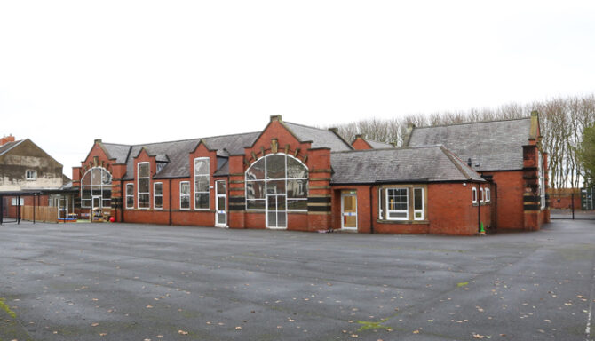 Brick school building with tall arched windows and a large glass entrance, set on a spacious paved playground with a few leaves